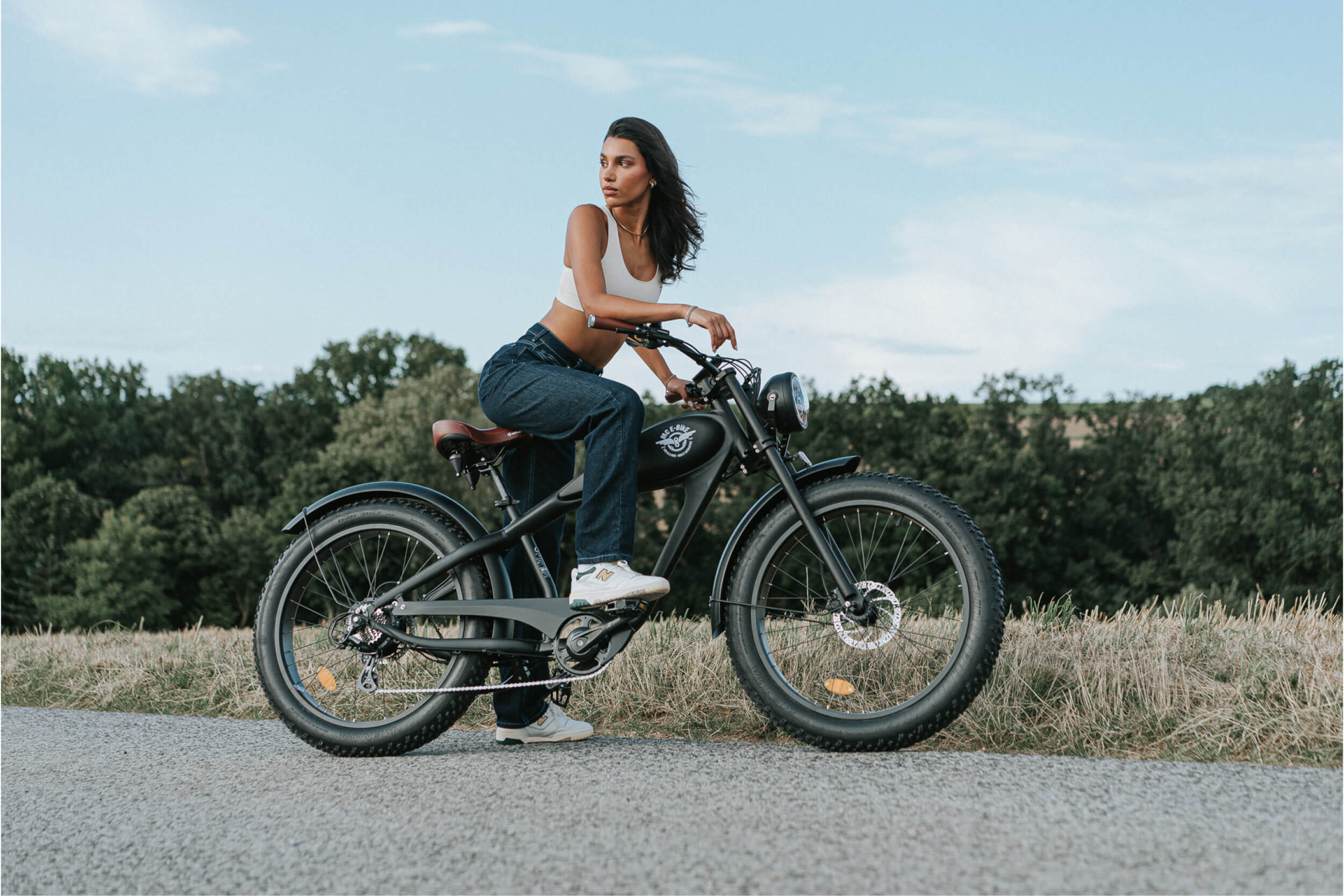 Lässiges E-Bike im Retro-Look: Junge Frau mit athlethischer Figur in Jeans und weißem Top, sich in der Gegend umschauend, in der Natur auf einem Fahrradweg