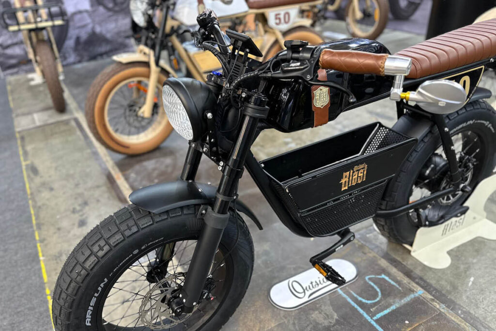Detailaufnahme eines schwarzen E-Bikes mit dem Namen MB Outsider 5.0 bei MC E-Bike, schwarzer Rahmen, brauner langezogener Sitz für zwei Personen und braune Griffe, Retro-Tank in Motorradoptik, schwarze Fatbike-Reifen, 20 Zoll