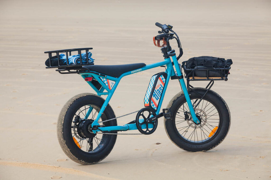 Aufnahme eines blauen E-Bikes am Strand, und den passenden Accessoires, mit dem Namen MB Ponyboy, hellblau-türkiser Rahmen, schwarzer langgezogener Sitz und schwarze Griffe, schwarze Fatbike-Reifen, 20 Zoll